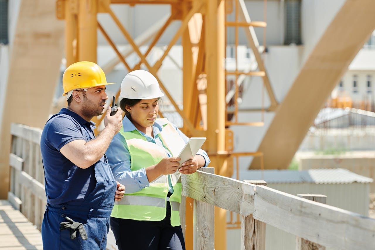 about-01 Two construction workers in hardhats discuss plans using a tablet and walkie talkie on-site.